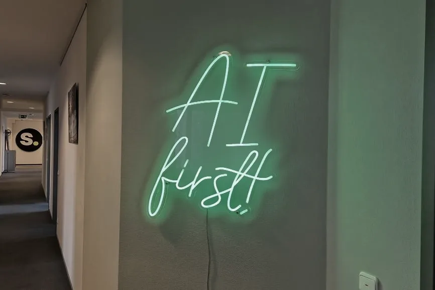 Grünes Neonschild mit dem Schriftzug AI first im Bürogang von schmidtmedia in Köln