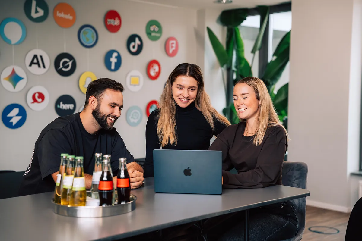 Team von schmidtmedia am MacBook vor der Wand mit Logos von Google, Meta, TikTok und KI-Plattformen im Kölner Büro